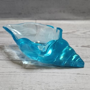 May include: A translucent, turquoise seashell-shaped bowl. The bowl has a wavy rim and a textured, ridged exterior, resembling a conch shell. The bowl is sitting on a light-colored wooden surface.