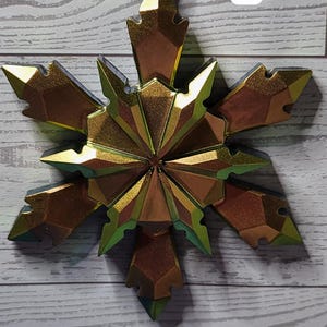 May include: A metallic gold snowflake ornament with a faceted design. The ornament is suspended by a silver string and has a brown loop at the top. A white rectangle with the number 7 is in the background.