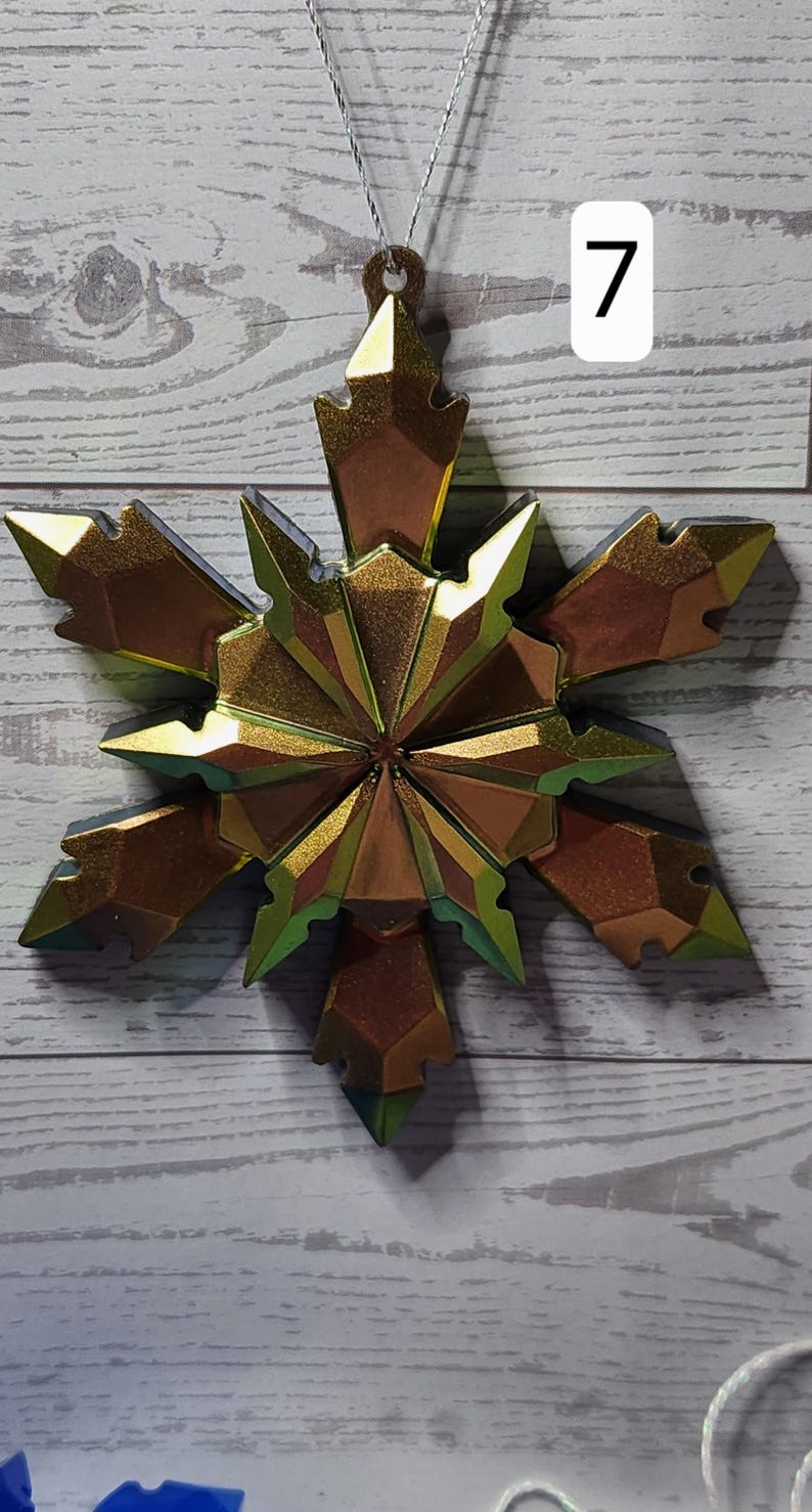 May include: A metallic gold snowflake ornament with a faceted design. The ornament is suspended by a silver string and has a brown loop at the top. A white rectangle with the number 7 is in the background.