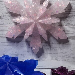 May include: A light pink snowflake ornament with glitter and the number 1. The snowflake is hanging from a white string against a wooden background. Other snowflake ornaments in blue and purple are visible.