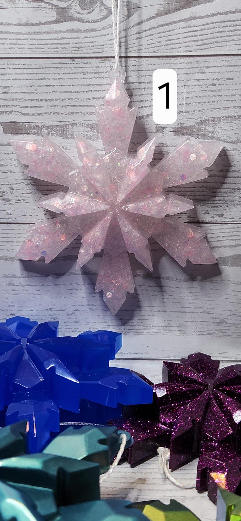May include: A light pink snowflake ornament with glitter and the number 1. The snowflake is hanging from a white string against a wooden background. Other snowflake ornaments in blue and purple are visible.