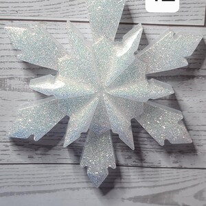 May include: A large, glittery snowflake ornament with a silver string for hanging. The snowflake is white with iridescent glitter, and has a number 12 in a white box. The ornament is set against a light wood-grain background.