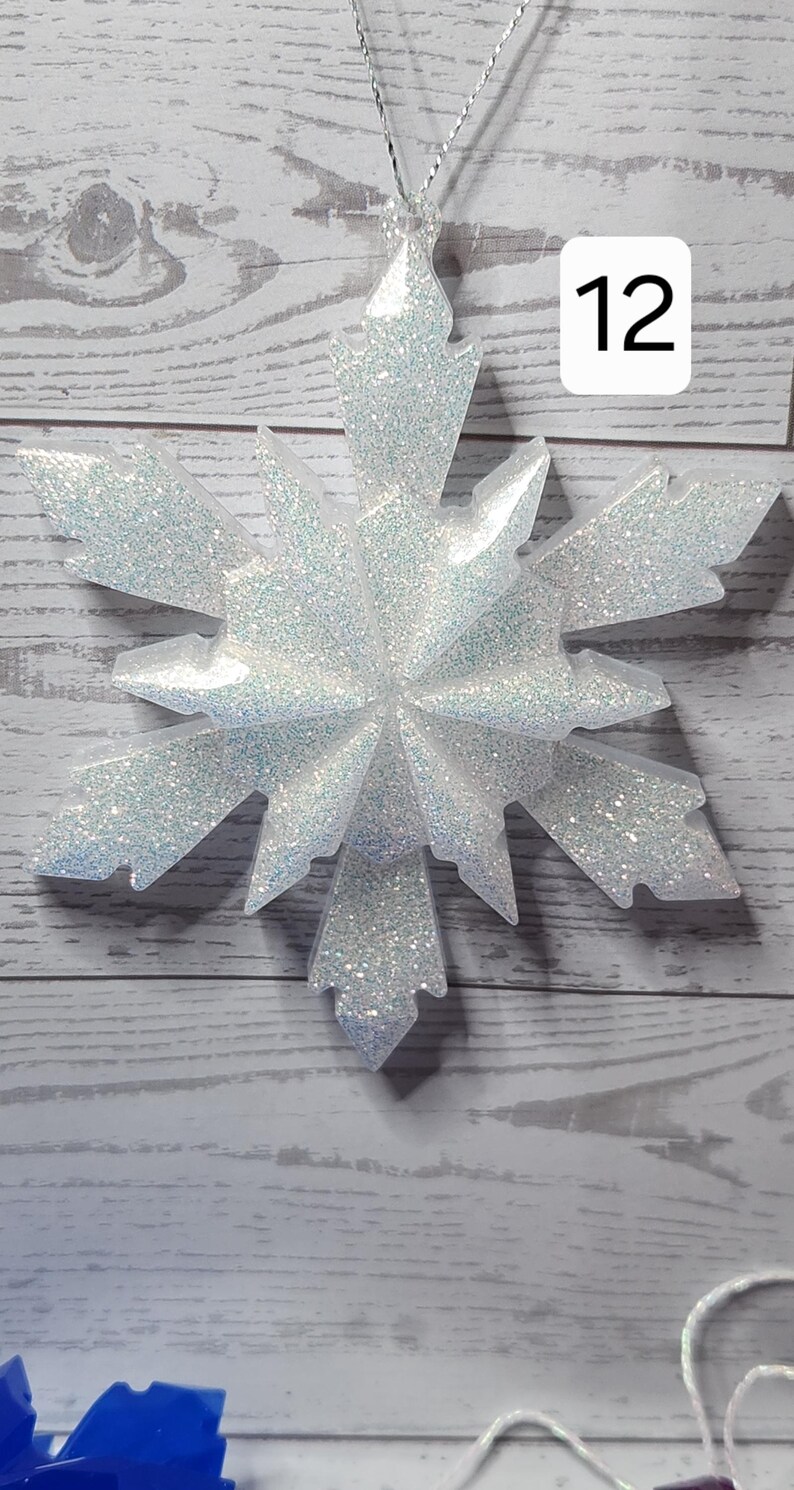 May include: A large, glittery snowflake ornament with a silver string for hanging. The snowflake is white with iridescent glitter, and has a number 12 in a white box. The ornament is set against a light wood-grain background.