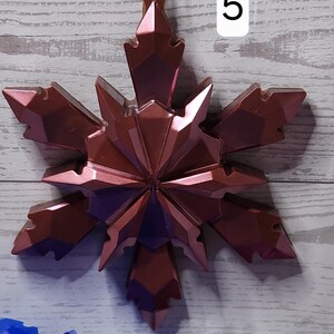 May include: A metallic rose-colored snowflake ornament with a faceted design, hanging by a white string. The number "5" is visible in the background. Other snowflake ornaments are visible in the background.