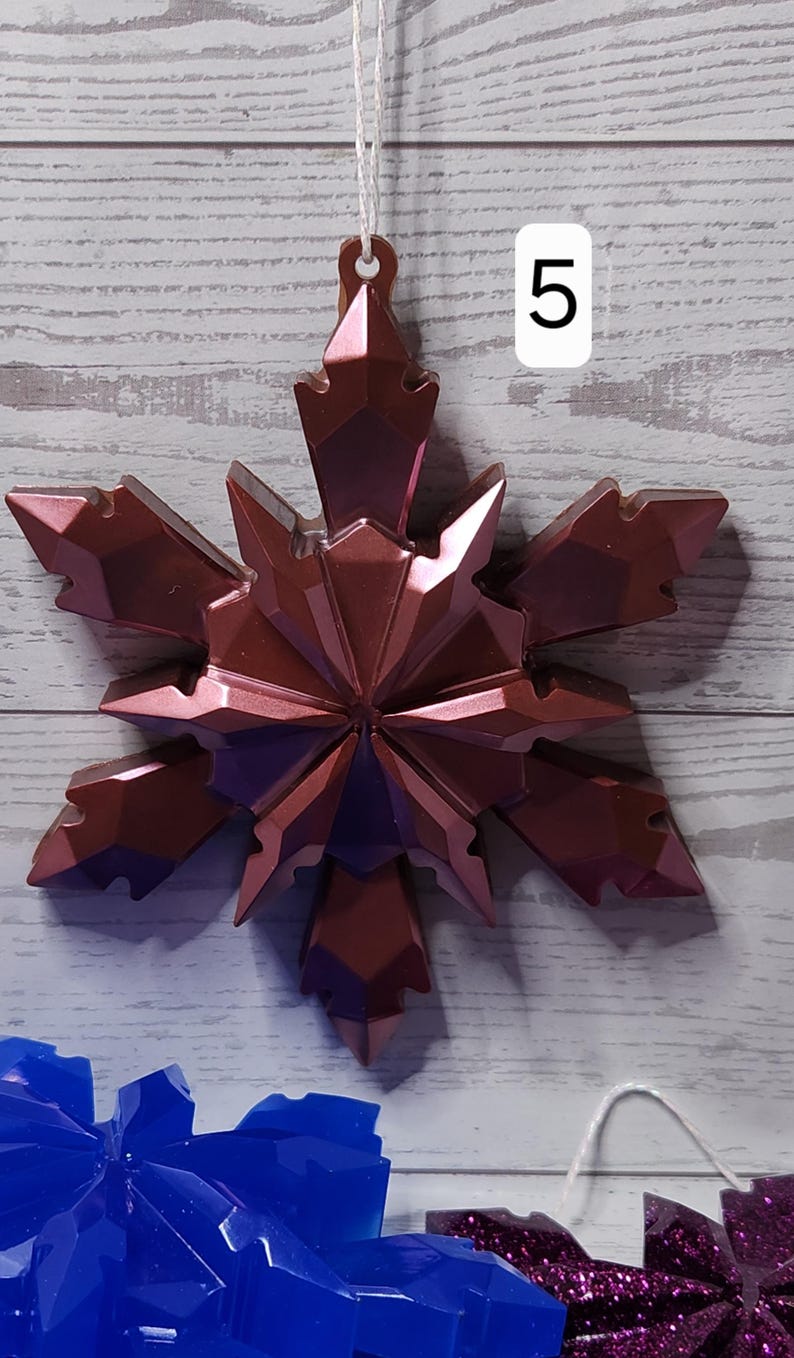 May include: A metallic rose-colored snowflake ornament with a faceted design, hanging by a white string. The number "5" is visible in the background. Other snowflake ornaments are visible in the background.