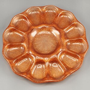May include: A glittery, orange-bronze resin paint palette with a central well and twelve surrounding wells. The palette has a rounded, petal-like shape, and the surface sparkles with glitter.