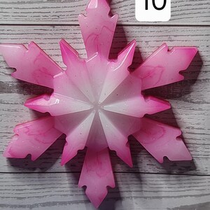 May include: Pink and white snowflake ornament with a glittery finish. The ornament has a starburst design and is hanging by a white string. The number "10" is in a white box in the upper right corner. The background is a light gray wood.