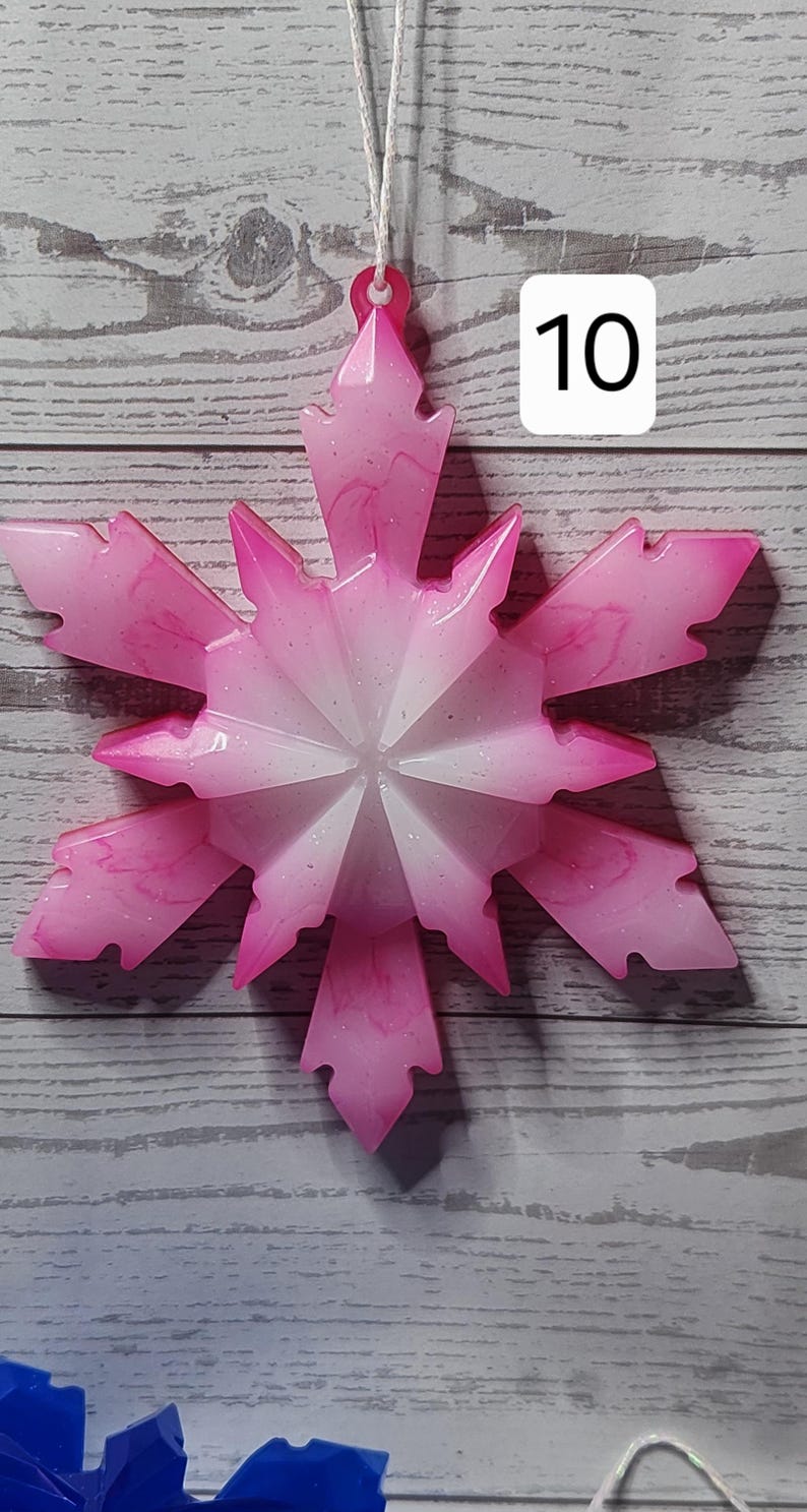 May include: Pink and white snowflake ornament with a glittery finish. The ornament has a starburst design and is hanging by a white string. The number "10" is in a white box in the upper right corner. The background is a light gray wood.