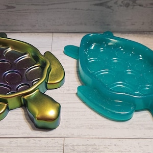 May include: Two turtle-shaped decorative trays. One is iridescent with green, gold, and purple hues. The other is a translucent teal color. Both have a textured interior and are displayed on a light wood surface.