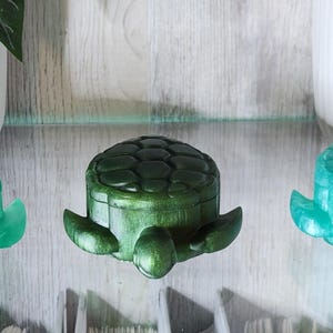 May include: Three decorative turtle-shaped boxes in shades of green. The boxes have a textured shell design and flipper-like appendages. The boxes are displayed on a glass shelf, with white planters in the background.