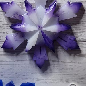 May include: A snowflake ornament with a purple and white design, hanging by a white string. The number 4 is in a white rectangle in the background. Other snowflake ornaments are visible in blue and purple.