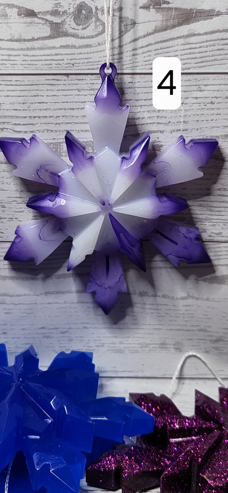 May include: A snowflake ornament with a purple and white design, hanging by a white string. The number 4 is in a white rectangle in the background. Other snowflake ornaments are visible in blue and purple.