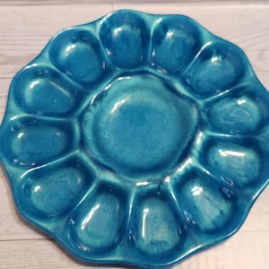 May include: A turquoise ceramic oyster serving platter with 12 individual compartments surrounding a central well. The platter has a scalloped edge and a glossy finish, perfect for serving appetizers or seafood.