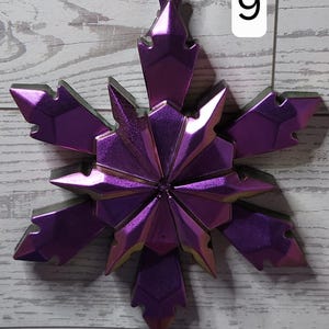 May include: A metallic purple snowflake ornament with a silver string for hanging. The snowflake has a geometric design with pointed edges and a reflective surface. A white number 9 is in the background.