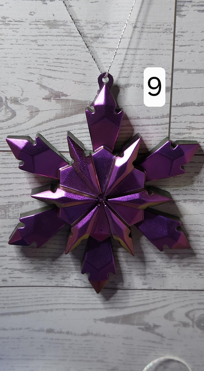 May include: A metallic purple snowflake ornament with a silver string for hanging. The snowflake has a geometric design with pointed edges and a reflective surface. A white number 9 is in the background.