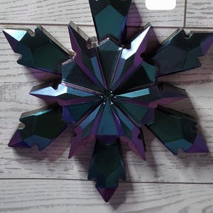 May include: Iridescent snowflake ornament with a faceted design. The ornament is primarily teal and purple, with a metallic sheen. The number "11" is displayed in a white box. The ornament is suspended by a silver string.