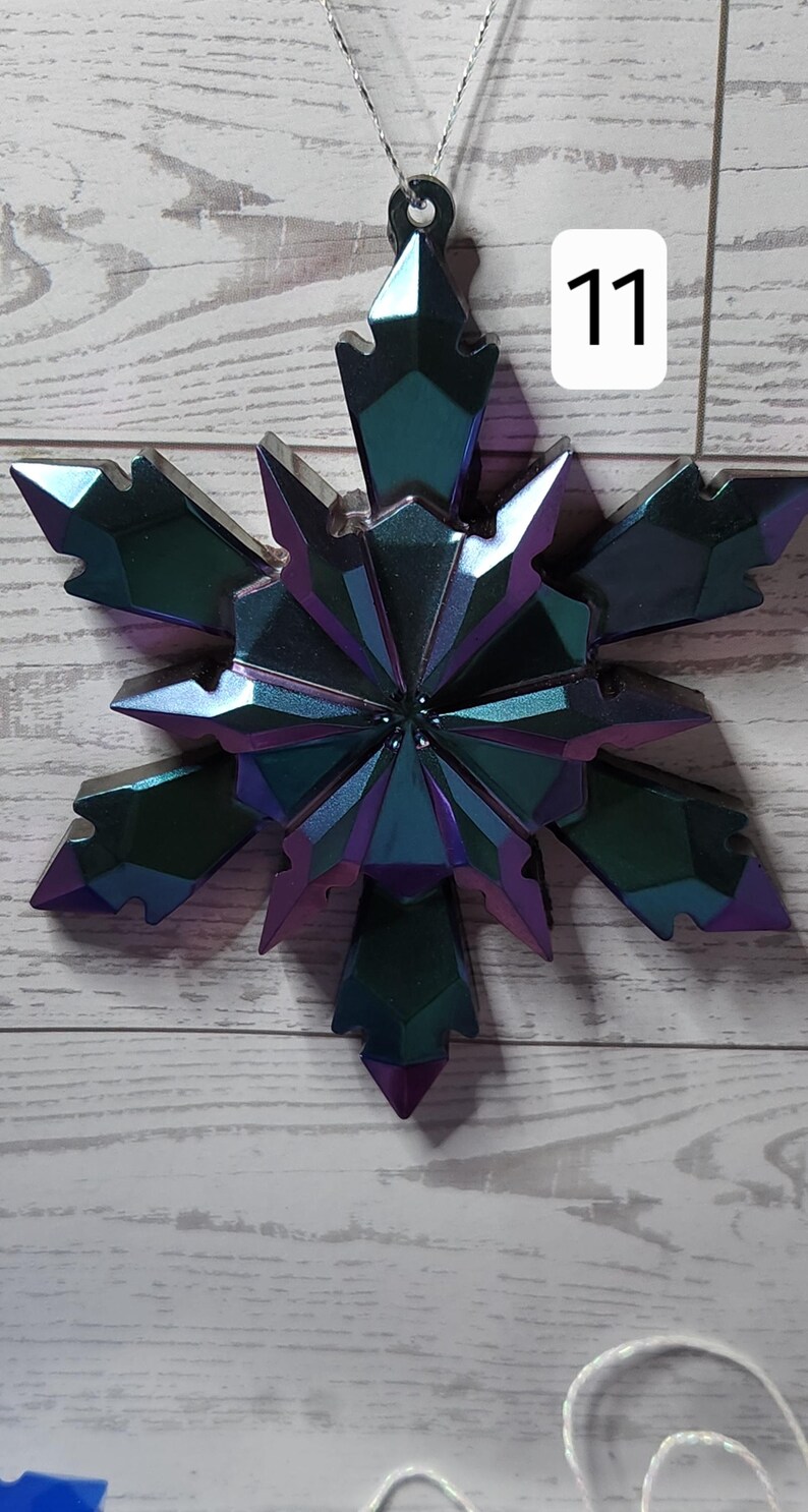May include: Iridescent snowflake ornament with a faceted design. The ornament is primarily teal and purple, with a metallic sheen. The number "11" is displayed in a white box. The ornament is suspended by a silver string.