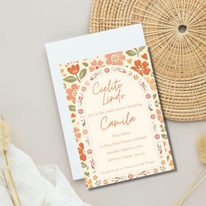 Op de afbeelding: Een rechthoekige uitnodigingskaart voor een babyshower met een cr&egrave;mekleurig boogframe en oranje en groene bloemmotieven. De tekst luidt: Cielito Lindo, Join us for a baby shower honoring Camila, Pizza Parlor, 111 Patty Place Denver, Colorado, Saturday August 14, 2025, RSVP to Tammy 555-555, Registered at Amazon and Target.