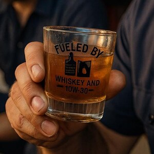 May include: A clear shot glass filled with amber liquid, labeled "Fueled By Whiskey and 10W-30." The glass is held by a person wearing a navy blue shirt and a cap. Another person is visible in the background.