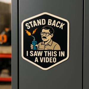 以下が含まれることがあります： 「STAND BACK I SAW THIS IN A VIDEO」のテキストが入った、黒とクリーム色のステッカー。メガネと口ひげをした漫画の男性が、オレンジ色の炎を上げたトーチを持っています。