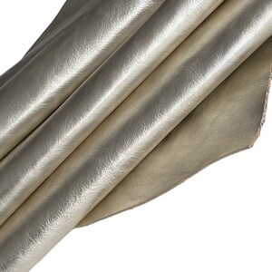 May include: A close-up of a folded piece of metallic gold leather. The leather has a textured surface and is folded in multiple layers, creating shadows and highlights. The edges of the leather are visible, showing the natural texture.