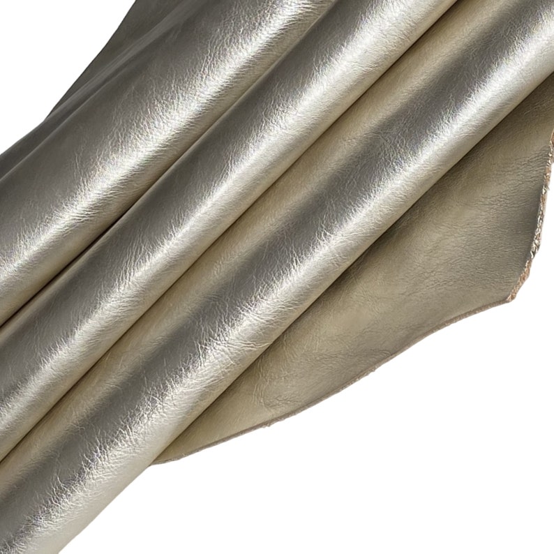 May include: A close-up of a folded piece of metallic gold leather. The leather has a textured surface and is folded in multiple layers, creating shadows and highlights. The edges of the leather are visible, showing the natural texture.