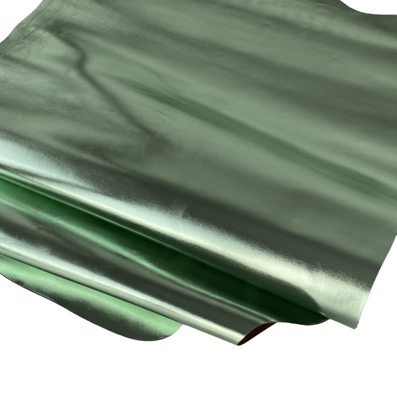 May include: A sheet of metallic green leather with a glossy finish. The leather is folded over itself, revealing a glimpse of the underside. The surface reflects light, creating highlights and shadows.