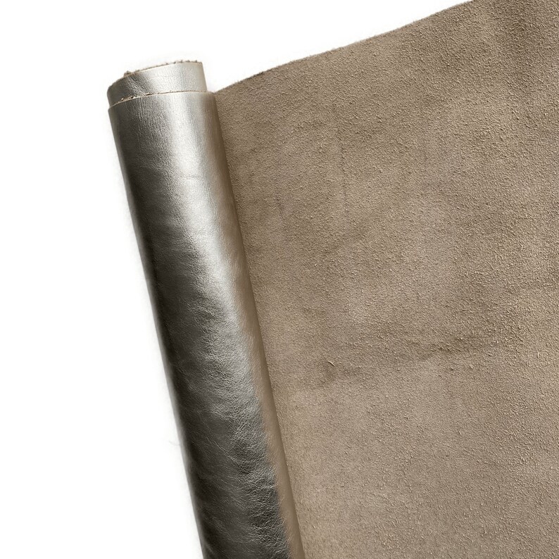 May include: A roll of metallic silver leather with a smooth, shiny surface. The unrolled portion reveals a tan suede-like texture. The leather is likely used for crafting or upholstery projects.