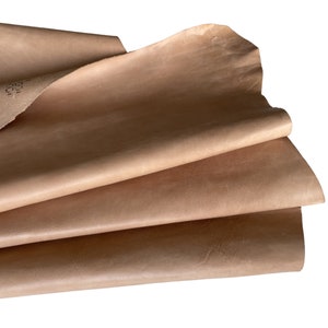 May include: Close-up of several sheets of tan-colored leather. The leather has a smooth, slightly textured surface and appears to be stacked. The edges of the leather are visible, revealing the layers. The lighting highlights the natural variations in the material.