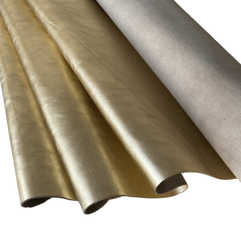 May include: Close-up of gold-colored leather sheets with a metallic sheen. The leather is folded, revealing a lighter, neutral-toned material on the reverse side. The texture appears smooth and supple, suitable for crafting.