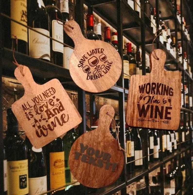 May include: Four wooden wine-themed serving boards with different sayings. The boards are hanging on a wall with wine bottles behind them. The sayings on the boards are "All you need is love and wine", "Save water drink wine", "Working nine to wine", and "Your text!"