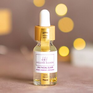 May include: A clear glass bottle with a gold label that reads "Helen's Hands 24k Facial Elixir Argan Rosehip Bergamot 100% Fresh". The bottle has a bamboo lid and contains gold flakes.