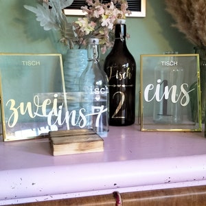Könnte beinhalten: Goldene und klare Glasrahmen mit den Wörtern "Tisch" und den Zahlen "eins", "zwei", "drei" und "fünf" in weißer Kalligraphie. Eine goldene Flasche mit dem Wort "Fisch" und der Zahl "2" in schwarzer Kalligraphie. Eine klare Glasflasche mit dem Wort "Tisch" und der Zahl "7" in schwarzer Kalligraphie.
