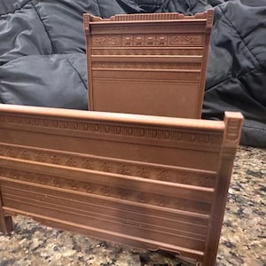 May include: Two brown miniature bed frames. The headboard and footboard are decorated with horizontal lines and floral patterns. The bed frames are made of a dark brown material and are likely part of a dollhouse or miniature set.