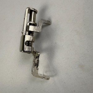 Könnte beinhalten: Ein Nähmaschinen-Nähfuß aus silberfarbenem Metall mit Federmechanismus und einer Basis aus transparentem Kunststoff. Das Metallteil ist mit der Gravur 161698 SINGER GT.BRIT versehen, welche den Hersteller und die Teilenummer angibt.