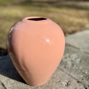 Puede incluir: Un jarrón de cerámica rosa claro con forma redondeada y bulbosa. El jarrón tiene un acabado liso y brillante.
