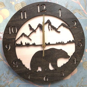 May include: A round wooden clock with a dark brown frame and a light-colored interior. The clock face features a bear, mountains, and trees. The clock hands are gold-colored, and the numbers are cut out of the wood.