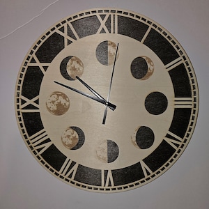May include: A round wooden clock with a light wood face and a dark wood outer ring. The clock features Roman numerals and phases of the moon. The clock hands are black.