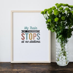 May include: A framed print with the text "My Train of Thought STOPS at no stations" in colorful letters. The print is on a wooden table next to a vase of green flowers.