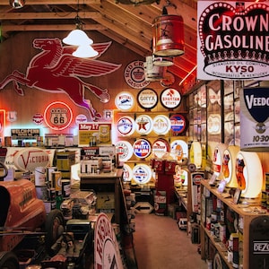 Op de afbeelding: Een verzameling vintage benzine- en olieborden en blikken in een garageomgeving. De borden tonen verschillende merken en logo's, waaronder Crown Gasoline, Veedol en Welch Penn. De borden zijn aan de muren en planken bevestigd en creëren een nostalgische sfeer.