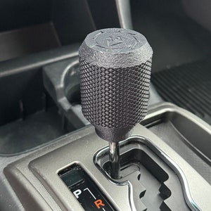 May include: A black gear shift knob with a textured surface and a raised triangle symbol on the top. The knob is attached to a silver gear shift lever with a black plastic housing. The gear shift lever is in a car with a silver interior. The gear shift lever has markings for P, R, N, 4D, 3, and 2L.