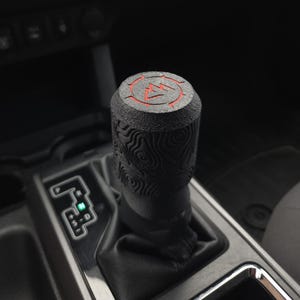 May include: A black gear shift knob with a textured wood grain pattern and a red mountain symbol on top. The knob is installed in a vehicle's interior, with the gear selector visible.