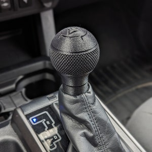 May include: A black gear shift knob with a textured surface and a raised design. The knob is attached to a black leather boot and is installed in a car's center console.