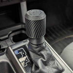 May include: A black gear shift knob with a textured, spiral design. The knob is installed in a car's gear shift lever.