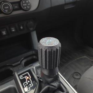 May include: A black gear shift knob with a textured, ridged design. The top of the knob features a circular emblem with a blue illuminated symbol. The shift lever is surrounded by a black leather boot, set in a vehicle's interior.