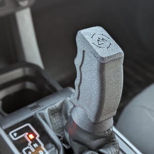 May include: A gray textured gear shift knob with a black leather boot. The knob has a raised logo with a mountain design. The gear shift is in the "P" position.