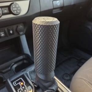 May include: A black, textured shift knob with a diamond pattern, installed in a vehicle. The knob is cylindrical and appears to be made of a durable material. The gear shift is surrounded by a black leather boot.