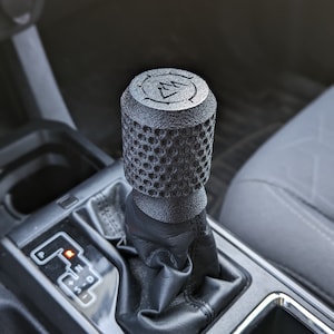 May include: Black gear shift knob with a textured surface and a triangular logo on top. The knob is installed in a car's gear shift.