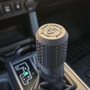 May include: A black gear shift knob with a textured diamond pattern and a mountain logo on top. The shift knob is part of a vehicle's interior, with the gear selector and other controls visible in the background.