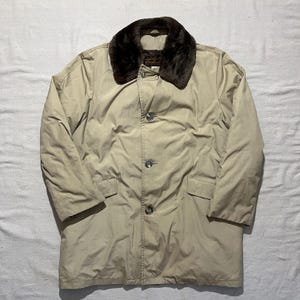 May include: A beige coat featuring a dark brown faux fur collar, a button-down front closure with three visible buttons, long sleeves, and two front flap pockets. The garment has a structured collar and a smooth, solid-colored outer fabric.